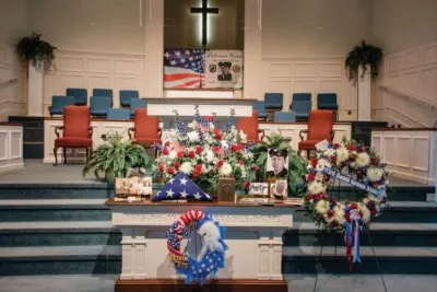 A church altar with a folded flag in a case, wreaths in red, white and blue, and a photograph of Weekley.