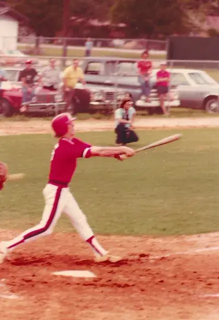 A batter in a red Devils uniform follows through on a swing at home plate, spectators and parked cars beyond the outfield fence.