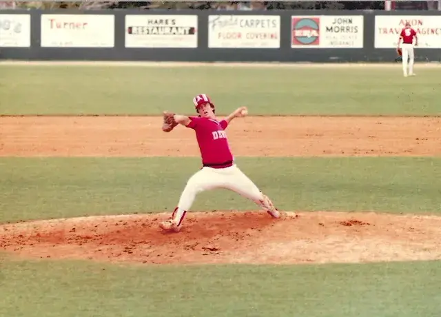 A pitcher in a red Devils uniform in mid-windup on the mound, the outfield fence lined with boards for Hare's Restaurant, Fred's Carpets and Avon Travel Agency.