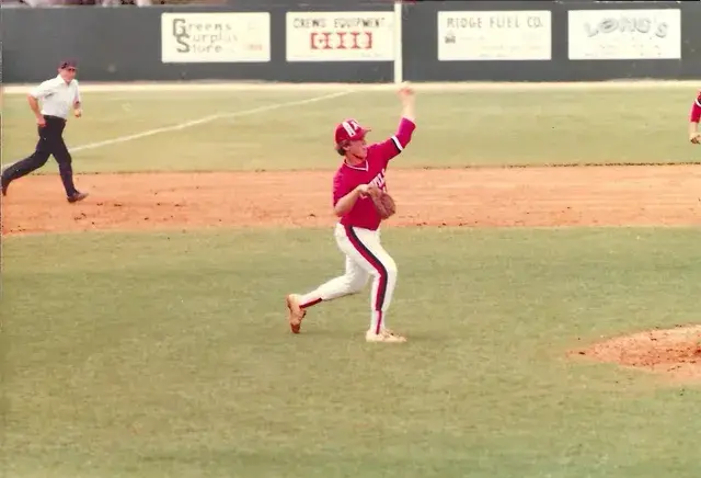 A red-uniformed player throws from the infield while an umpire runs behind him; boards for Greens Surplus Store, Ridge Fuel Co. and Long's line the fence.
