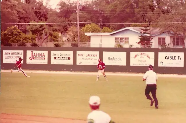 A wide view of the outfield with a fielder running toward a fence carrying boards for Jahna Concrete, Badcock and Cable TV, houses beyond.