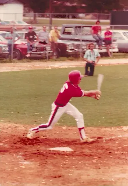A batter in a red uniform in mid-swing at the plate, spectators leaning on parked cars behind the backstop.