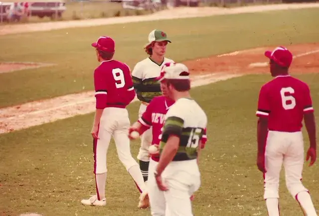 Four players meet in the infield: two in red Devils jerseys numbered 9 and 6, two in white jerseys with green stripes.