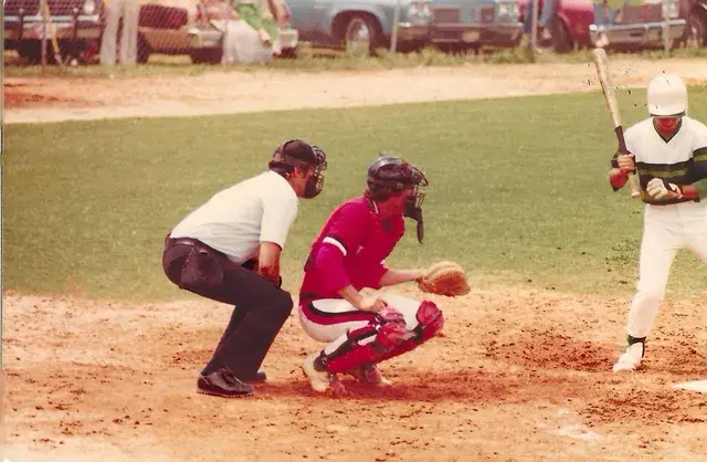 A catcher in red gear crouches behind the plate as the umpire leans in and a batter in a white-and-green jersey waits.