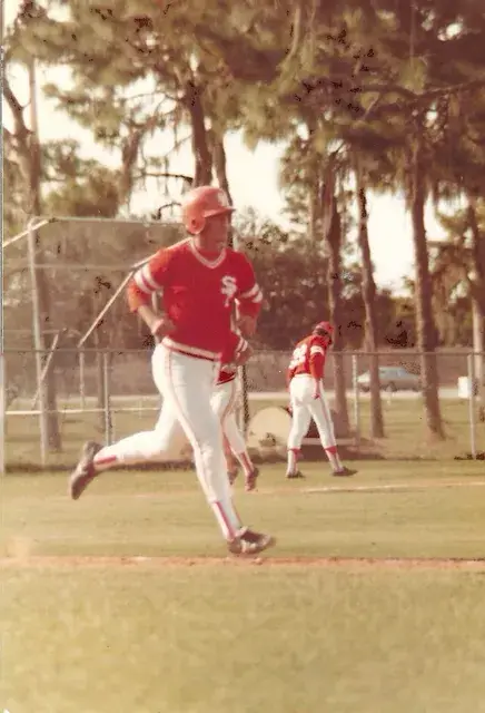 A runner in a red S jersey and batting helmet rounds the bases past a chain-link fence.