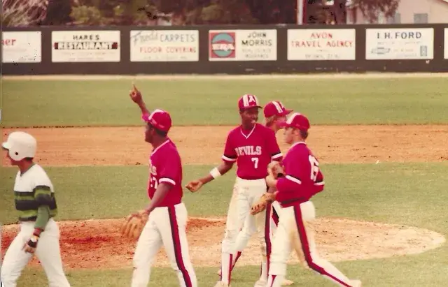 Three players in red Devils jerseys, one pointing skyward, celebrate near the mound as an opponent in green stripes walks past.