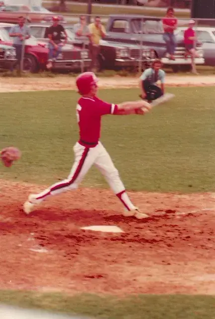 A batter in a red uniform swings at home plate with a row of parked cars behind the fence.