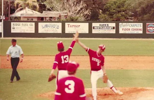 A red-uniformed pitcher, number 5, delivers while infielders 13 and 3 look on and an umpire watches from the base line.