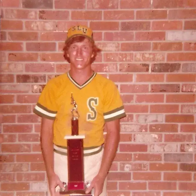 A young man in a gold and green S jersey and cap holds a trophy in front of a brick wall.