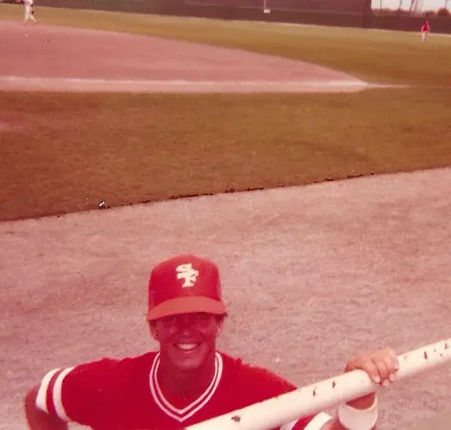 A smiling player in a red cap and jersey leans on a rail beside the field.