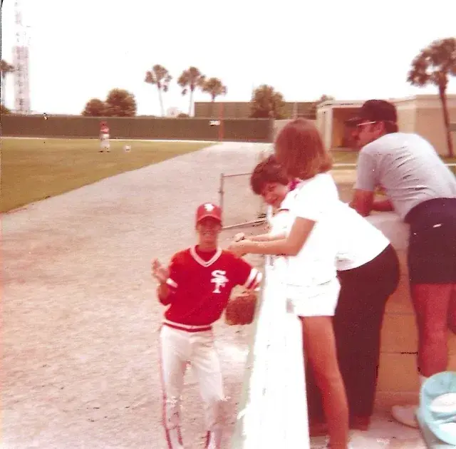 A player in a red SF jersey stands at a fence talking with spectators, palms and the outfield behind.