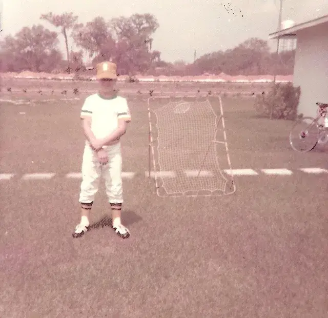 A boy in a baseball uniform and cap stands on a lawn beside a pitch-back practice net, a bicycle behind him.
