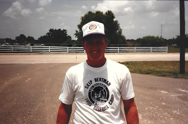 A man in a cap and a 1985 LSU Skip Bertman Baseball Camp T-shirt smiles beside a white fence.