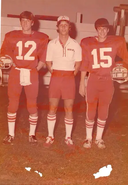 Three young men in red football-style jerseys numbered 12 and 15 with a coach between them, a faded colour print.