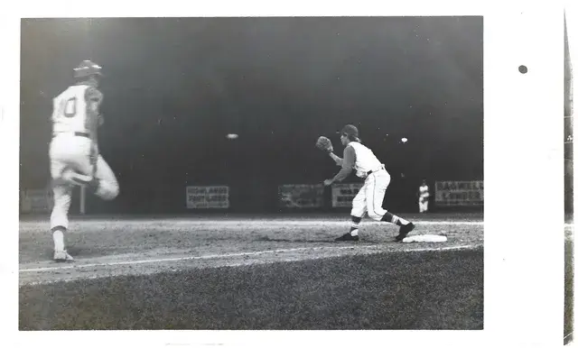 A black-and-white night photograph of a pitcher in full leg kick and an infielder set at first base under the lights.
