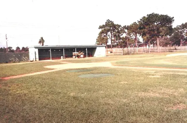 An empty baseball diamond with a covered dugout and pine trees behind it.