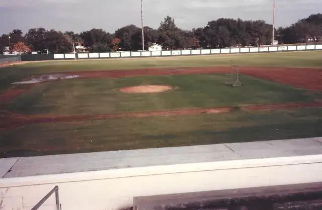 A wet infield seen from the stands: mound, base paths, a batting cage and a white outfield fence.