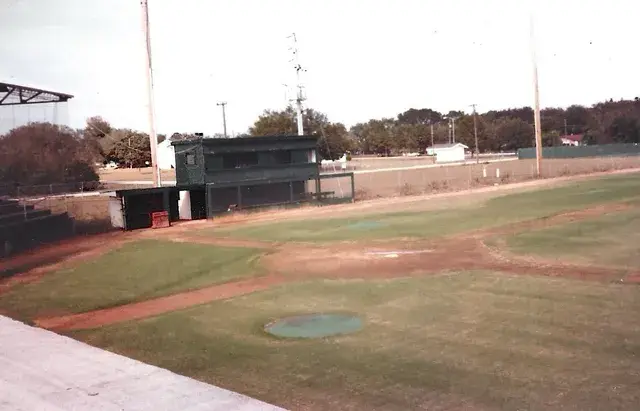 A covered dugout and a two-storey press box beside the diamond under light poles.