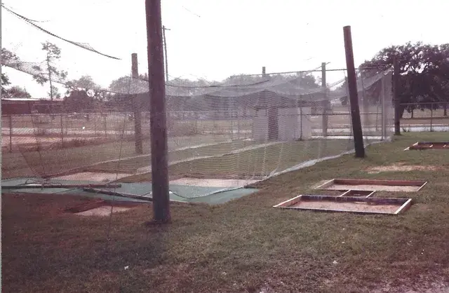 Batting cages with netting and pitching mats on the grass beside a field.