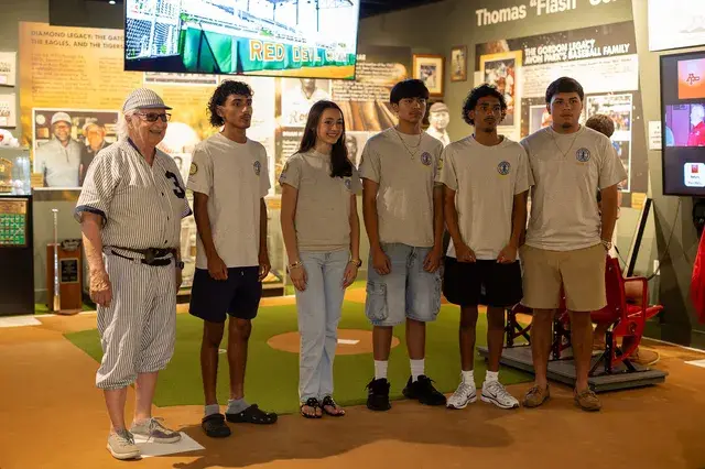 The man in the vintage pinstriped number 3 uniform with five young volunteers in grey shirts on the gallery turf.