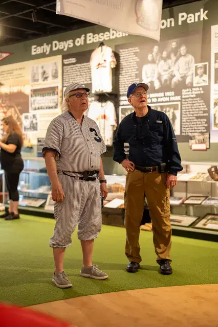 The man in the vintage pinstriped number 3 uniform with a visitor in front of the Early Days panel.