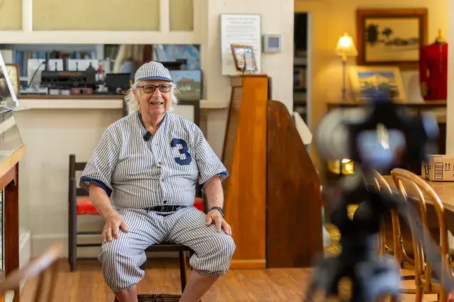 The man in the vintage pinstriped number 3 uniform seated in the Depot office, a camera in the foreground.