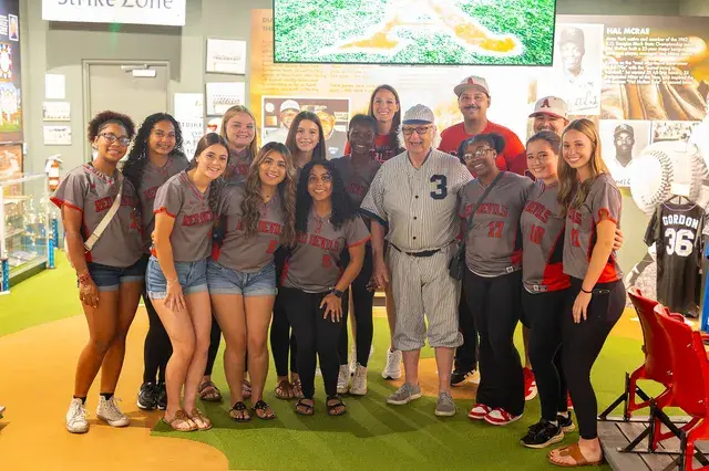 A girls' softball team in grey jerseys pose with the man in the vintage pinstriped number 3 uniform beneath a Strike Zone sign.