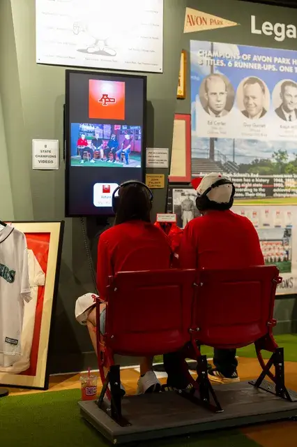 Two people sit in red stadium seats watching a screen.