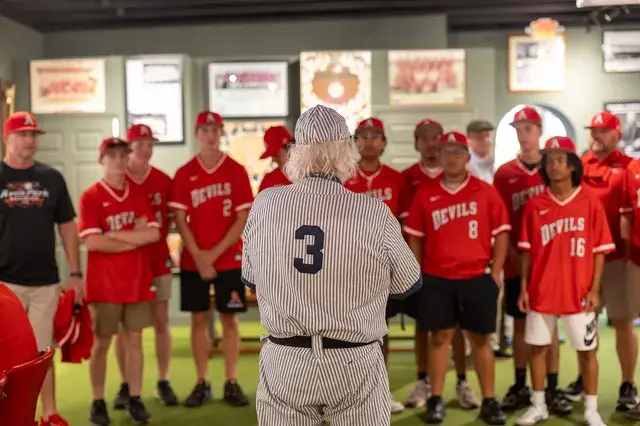 The man in the vintage pinstriped number 3 uniform addresses the Devils baseball team in red jerseys.
