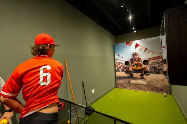 A player in a red number 6 jersey at the pitching game, a catcher target painted on the wall.
