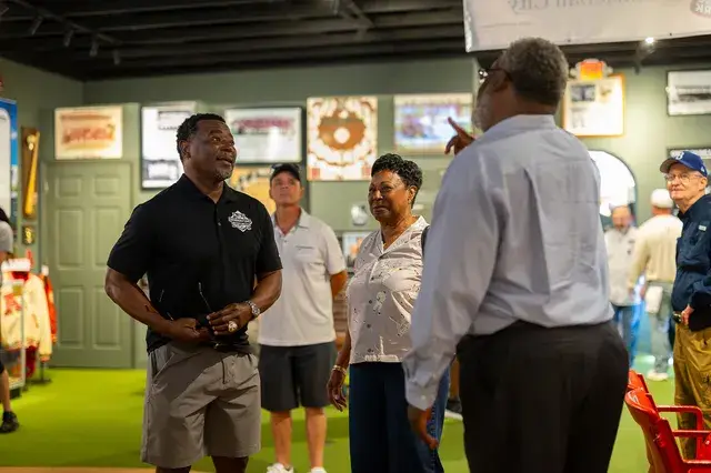 A man in a black polo talks with guests in the gallery.