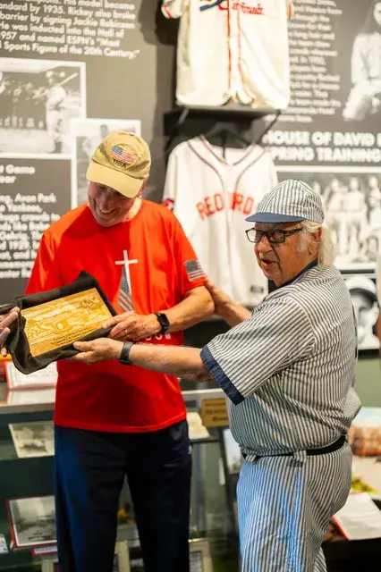 A man in a red shirt and the man in the vintage pinstriped number 3 uniform hold a framed plaque before the House of David panel.