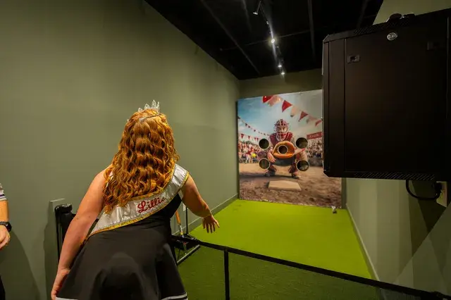 A young woman in a pageant sash throws at the Strike Zone pitching game.
