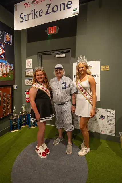 Two pageant winners in sashes pose with the man in the vintage pinstriped number 3 uniform under the Strike Zone sign.