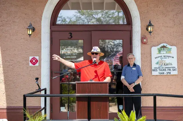 A man in a red shirt speaks at the podium, the Depot hours sign beside him.