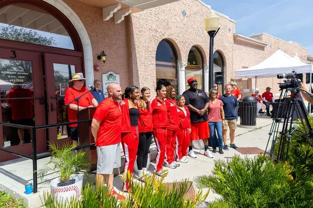 Team and guests in red along the porch railing, a camera crew beyond.