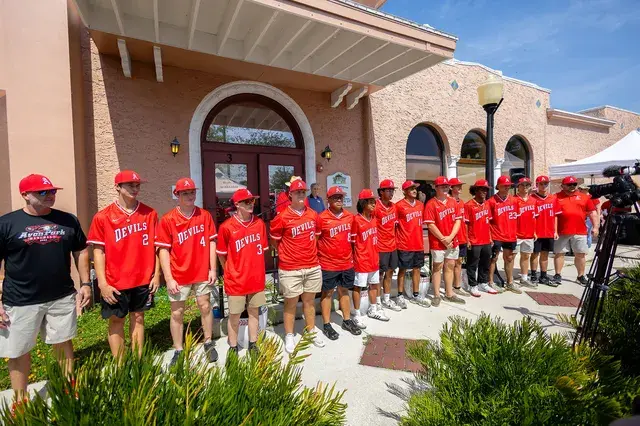 The Devils baseball team in red jerseys and caps lines up in front of the Depot.