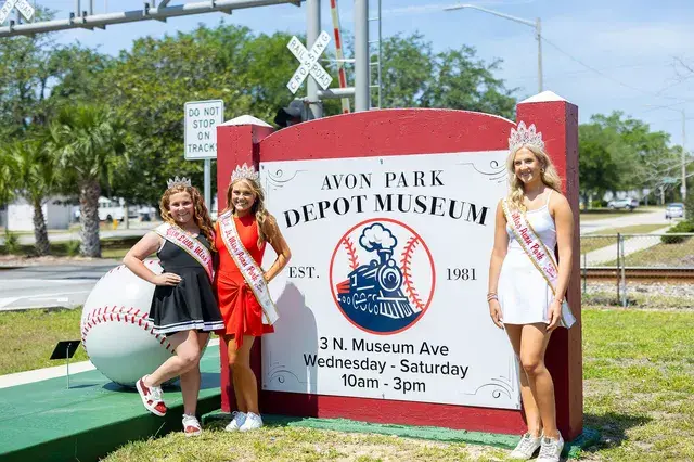 Pageant winners pose beside the Avon Park Depot Museum sign and a giant baseball.