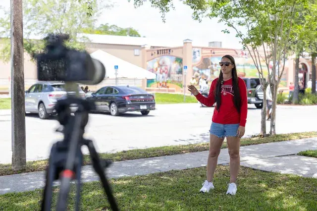 A woman in red addresses a video camera on the lawn.