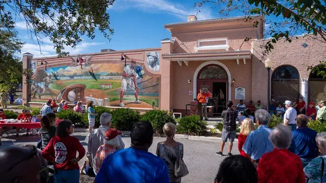 A crowd gathered on the lawn in front of the Depot Museum's new baseball mural under a blue sky.
