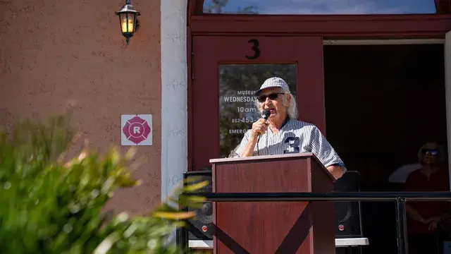 A man in a cap and pinstriped number 3 jersey speaks into a microphone at a podium beside the Depot's door.