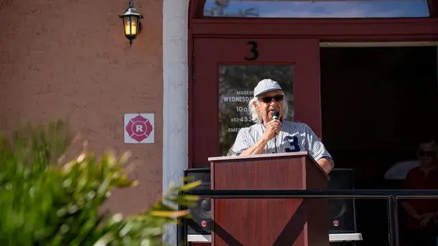 The same speaker in pinstripes at the podium, seen from the lawn through the shrubs.