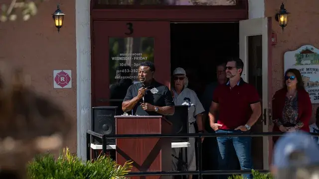 A speaker in a dark shirt at the podium with guests standing in the doorway behind him.