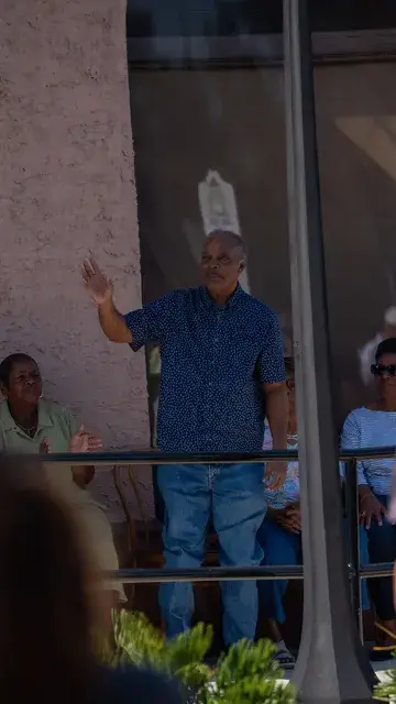 A man in a blue shirt raises a hand at the podium, seen through the porch railing.