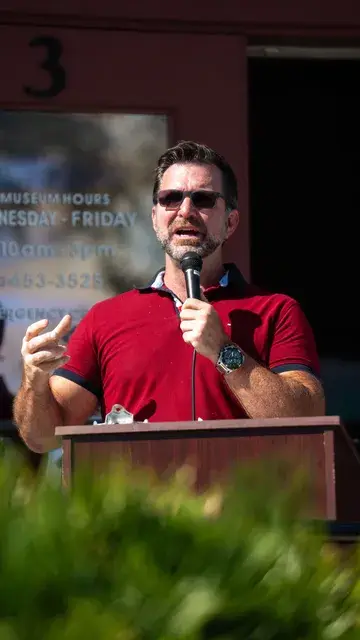 A man in a red polo and sunglasses speaks into a microphone at the podium.