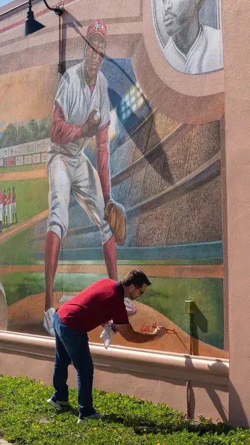 A man in a red polo crouches to look at the base of the mural's pitcher figure.