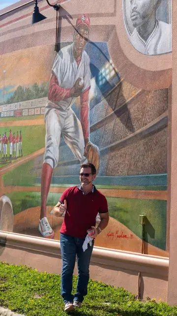 A man in a red polo stands before the mural's pitcher beneath the portrait medallion.