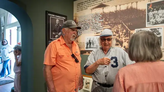 The man in the vintage pinstriped number 3 uniform talks with two guests, panels on the early days of Avon Park baseball behind them.