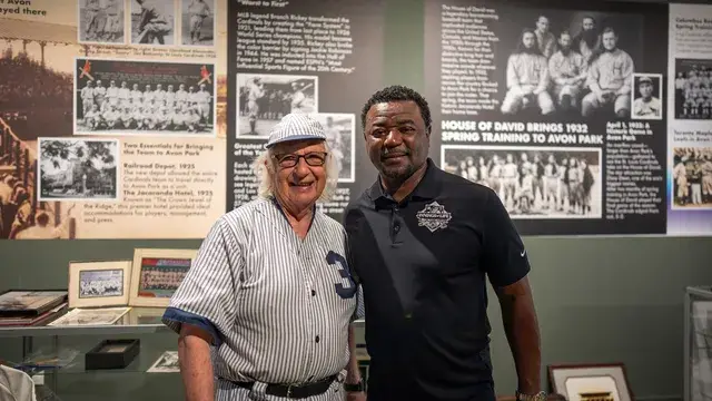 The man in the vintage pinstriped number 3 uniform and a man in a black polo pose by the wall of team photographs.