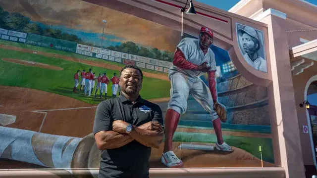 A man in a black polo stands with arms folded beneath the mural's pitcher.